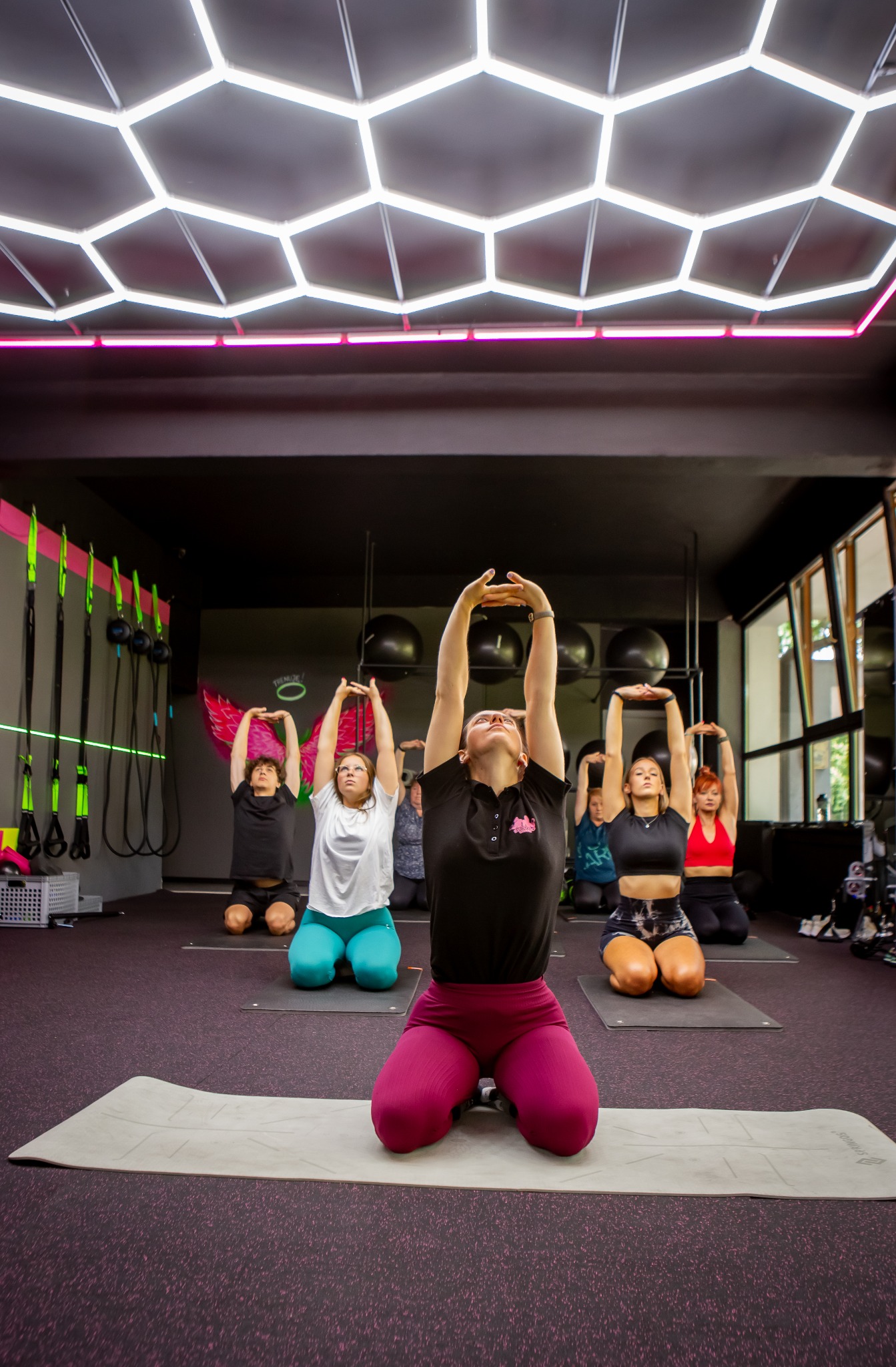 Grupa osób w sali fitness klęczy na matach, unosząc ręce nad głowami; w tle widoczne czarne piłki i grafika skrzydeł na ścianie.
