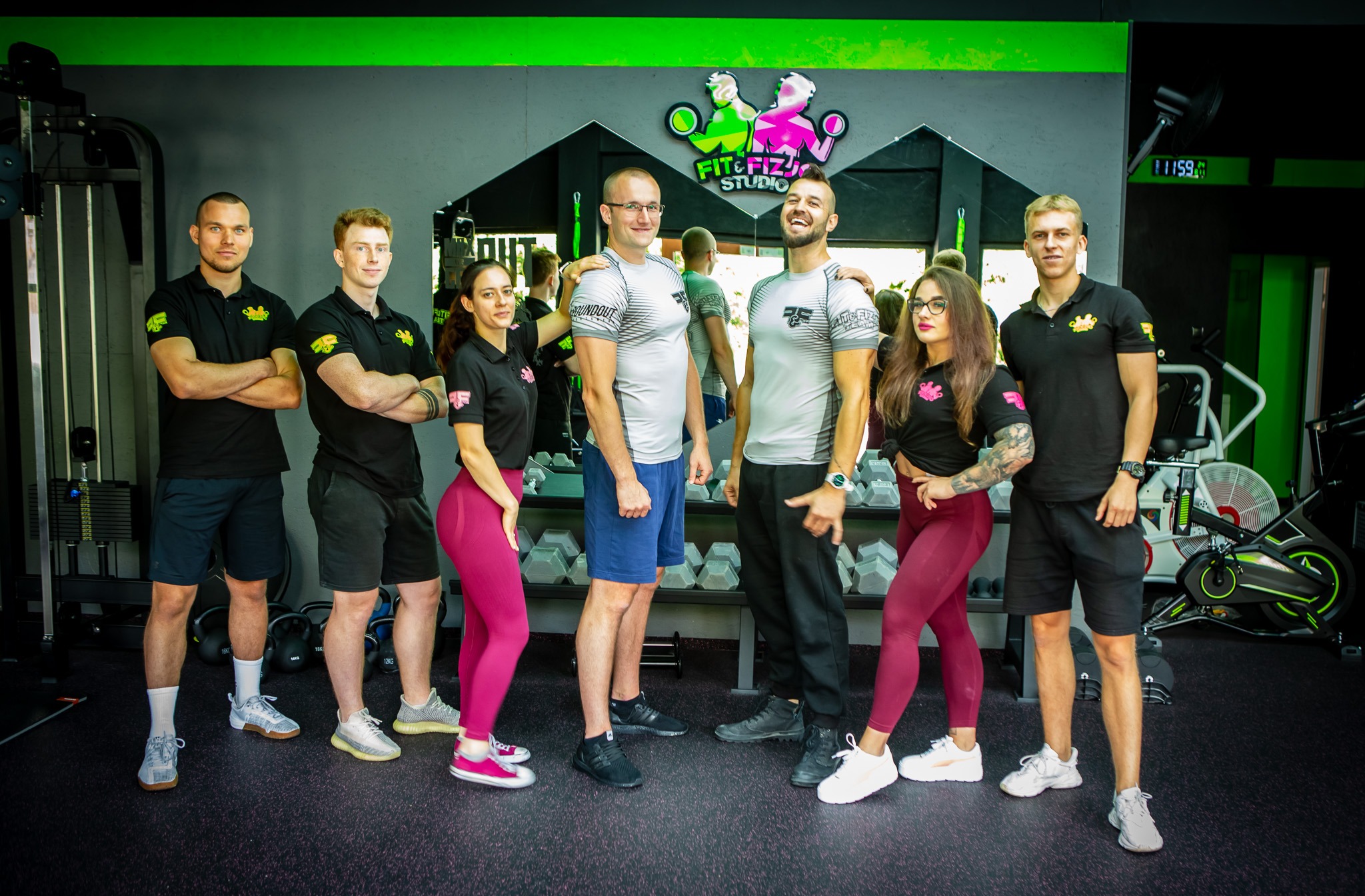 Grupa sześciu osób, w tym dwie kobiety, w strojach sportowych, pozujących w siłowni przed lustrem z widocznym logo Fit&Fiz Studio, obok sprzęt do ćwiczeń.