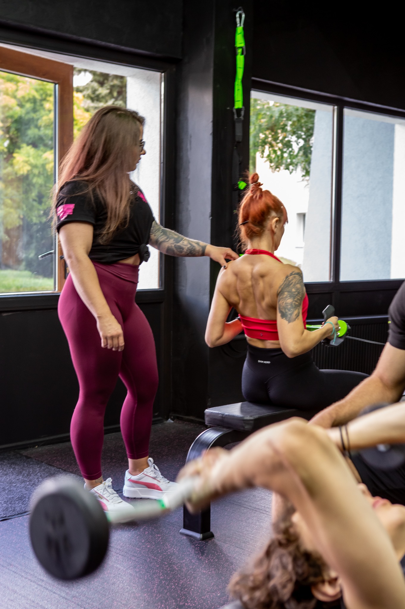 Kobieta w bordowych legginsach i czarnej koszulce z tatuażem na ramieniu obserwuje trening innej kobiety na siłowni, która siedzi na ławce i ćwiczy mięśnie pleców za pomocą zielonego urządzenia...