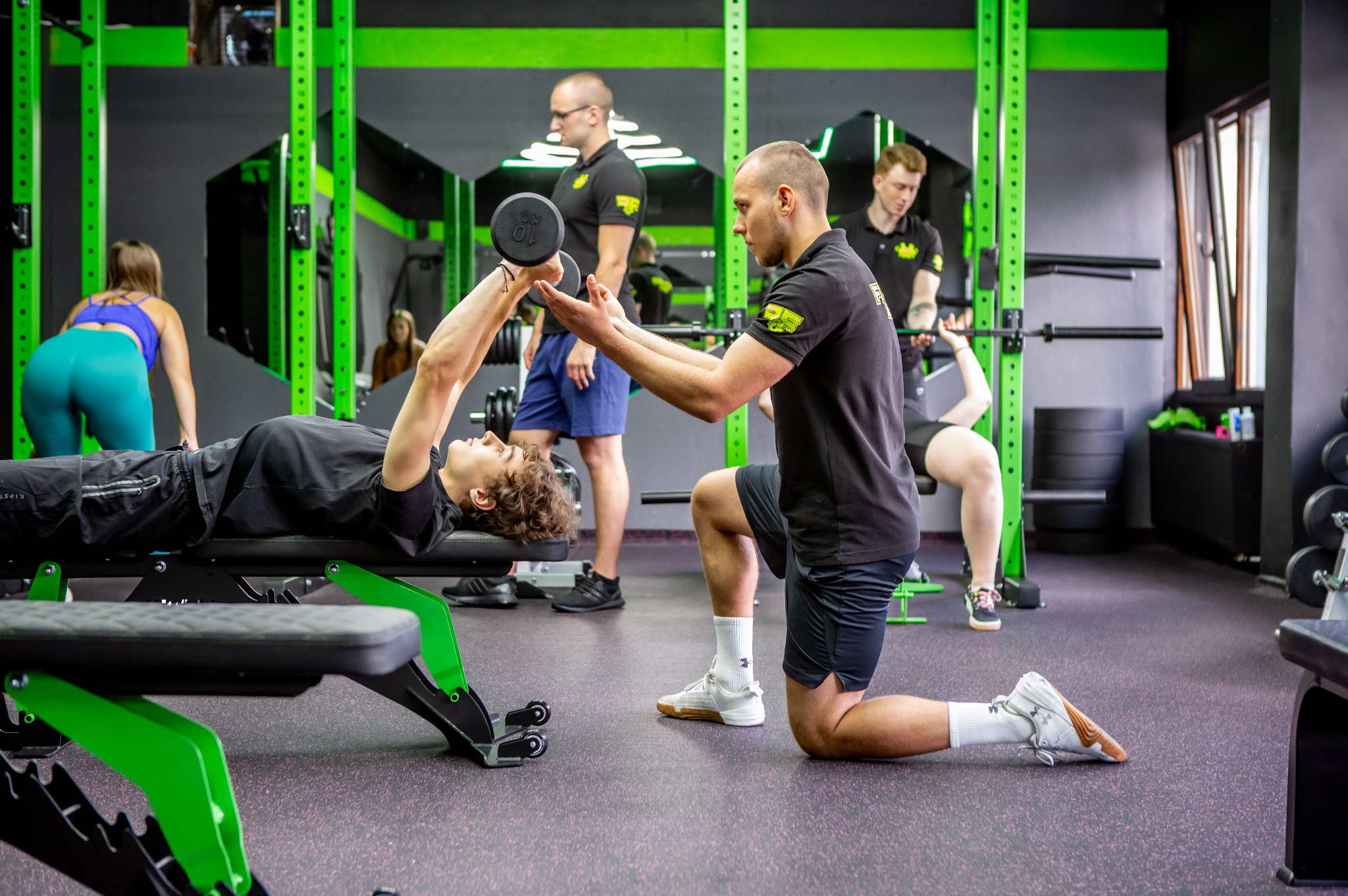 Mężczyzna asekuruje ćwiczącego z hantlami na ławce w siłowni, w tle inni ćwiczący i sprzęt do ćwiczeń, neonowe zielone akcenty.