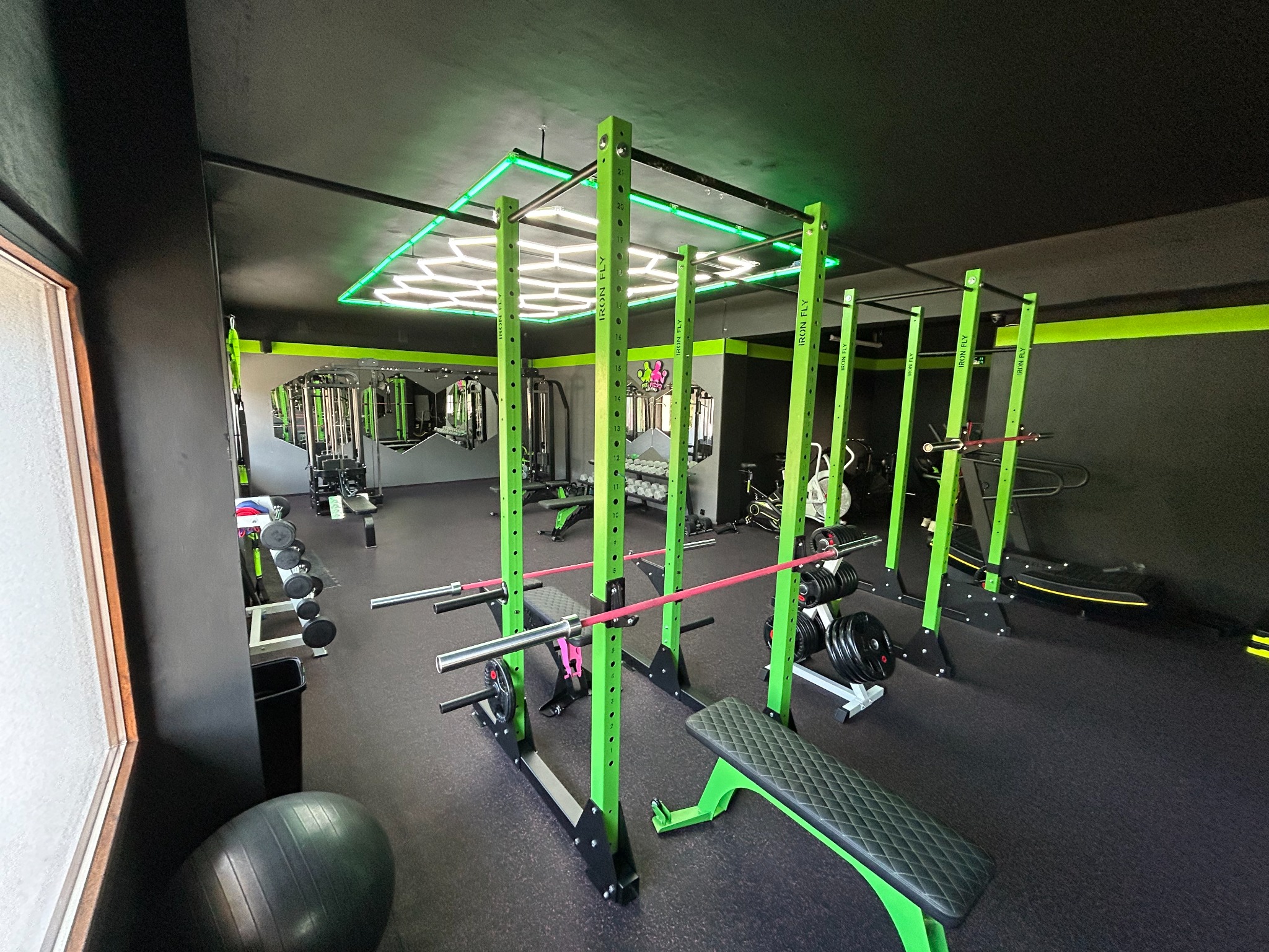Profesjonalnie wyposażona siłownia z zielonymi elementami sprzętu, oświetleniem LED i różowymi sztangami, widok na różne maszyny i hantle w tle.