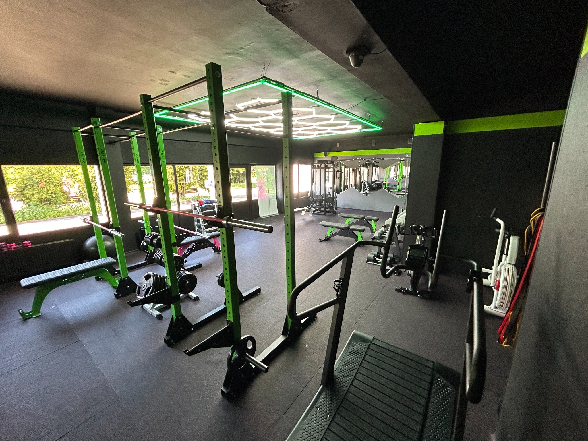 Wnętrze siłowni z wyposażeniem do ćwiczeń siłowych: klatka do ćwiczeń, ławeczki, bieżnia bez napędu, sztangi, obciążenia, oświetlenie LED.