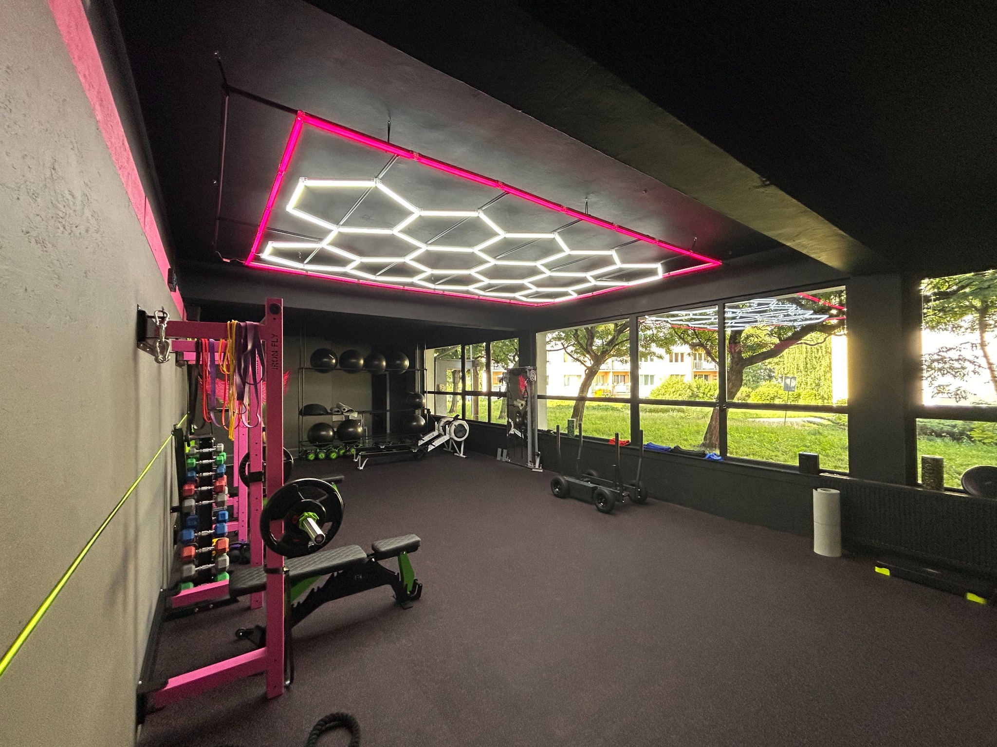 Nowoczesna siłownia z neonowym oświetleniem sufitowym w kształcie plastra miodu, sprzęt do ćwiczeń siłowych i cardio, widok na zieleń za oknem.
