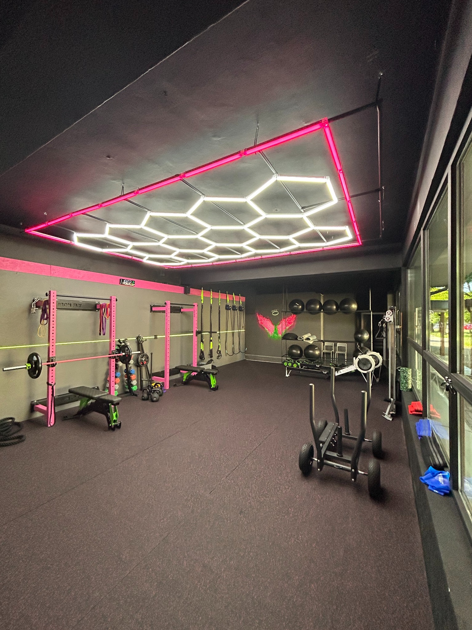 Nowoczesna siłownia z czarno-różowym wystrojem, wyposażona w sprzęt do ćwiczeń siłowych i cardio, oświetlona geometryczną lampą sufitową z różowym obramowaniem.