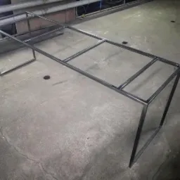 Surowy szkielet metalowej konstrukcji stołu, spawany z profili kwadratowych, leżący na szarej, betonowej posadzce w warsztacie.