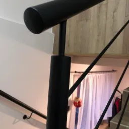 Czarna, metalowa konstrukcja schodów spiralnych z okrągłym przekrojem elementów, widoczna z dołu, z dekoracyjną bombką w tle.