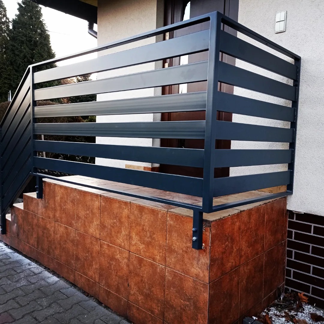 Nowoczesna, pozioma balustrada metalowa w kolorze grafitowym, zabezpieczająca schody wejściowe do domu, z widocznymi mocowaniami do podłoża wyłożonego brązowymi płytkami.
