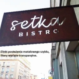 Rdzawy metalowy szyld z wyciętymi literami nazwy 'Setka Bistro', zawieszony przed budynkiem w miejskiej scenerii.