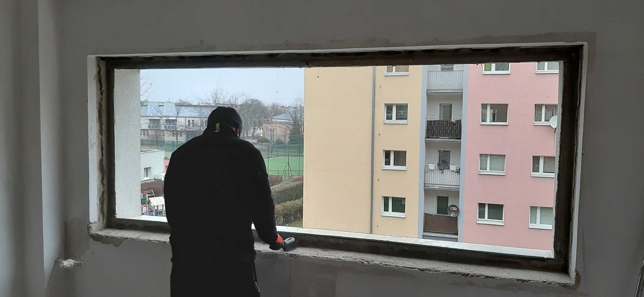 Mężczyzna w czarnej kurtce przygotowuje otwór okienny do montażu okna, widok na blok mieszkalny z zielonym boiskiem w tle.