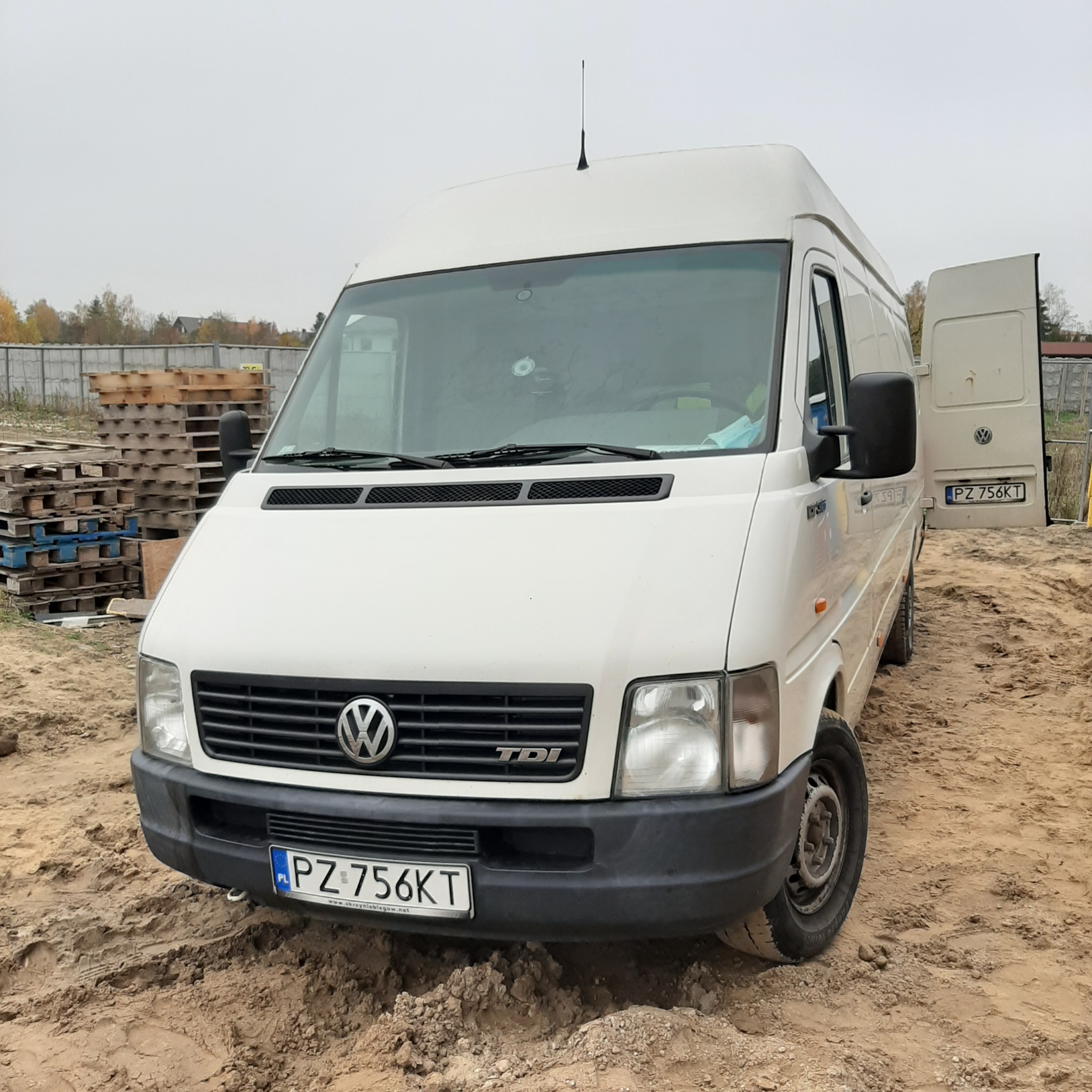 Biały samochód dostawczy Volkswagen LT z otwartymi drzwiami, zaparkowany na piaszczystym terenie z paletami w tle.