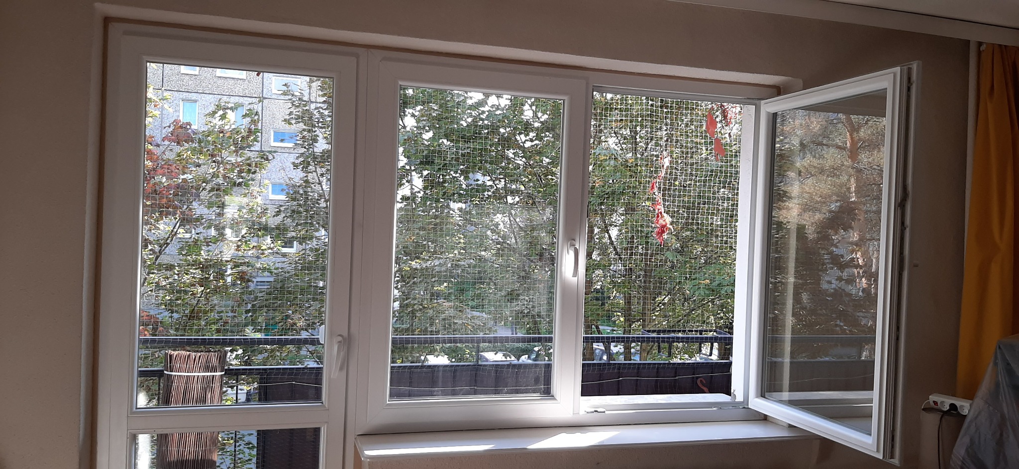 Białe okno trzyskrzydłowe z moskitierą, jedno skrzydło otwarte, widok na zieleń za oknem.