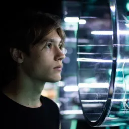 Młody mężczyzna w czarnym t-shircie patrzy z zaciekawieniem na rybę w akwarium z turkusowym oświetleniem.