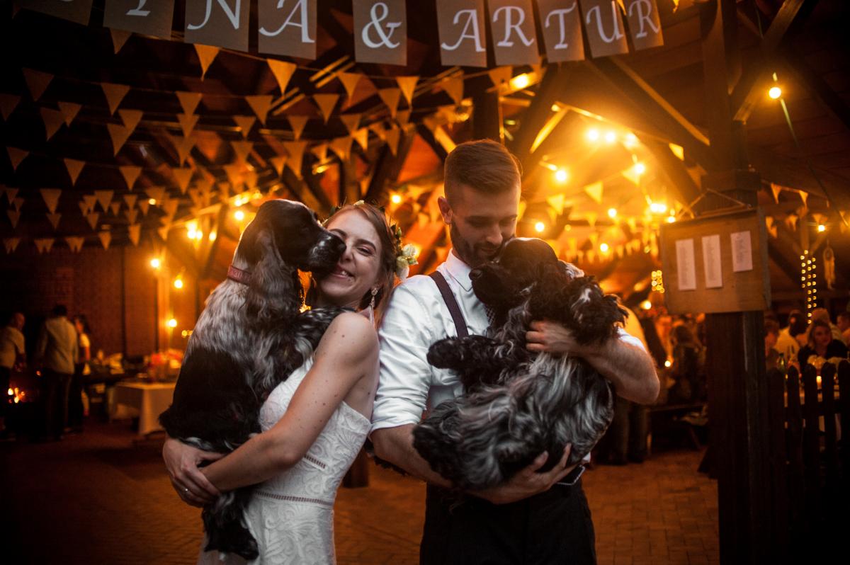 Para młoda trzymająca dwa psy rasy cocker spaniel na tle dekoracji weselnych z napisem 'Tina & Artur' i girlandami żarówek.