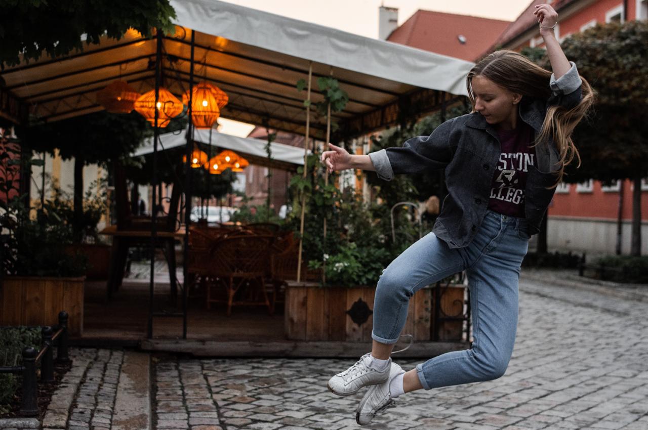Młoda kobieta w jeansowej kurtce i dżinsach skacze na brukowanej ulicy przed restauracją z oświetlonymi lampionami.