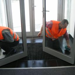 Delta Plus Sp. z o.o. Sprzątanie obiektów - Dwóch pracowników w pomarańczowych kamizelkach czyści podłogę przy szklanych drzwiach wejściowych, widoczna czarna mata podłogowa i ciemne płytki.
