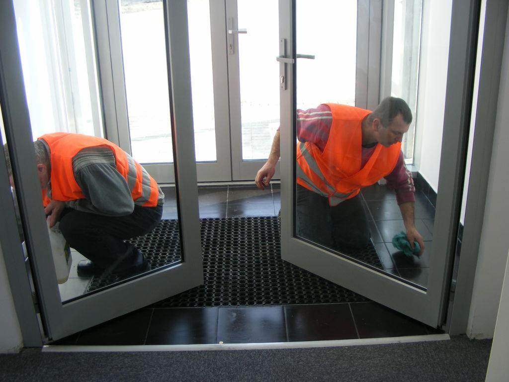 Dwóch pracowników w pomarańczowych kamizelkach czyści podłogę przy szklanych drzwiach wejściowych, widoczna czarna mata podłogowa i ciemne płytki.