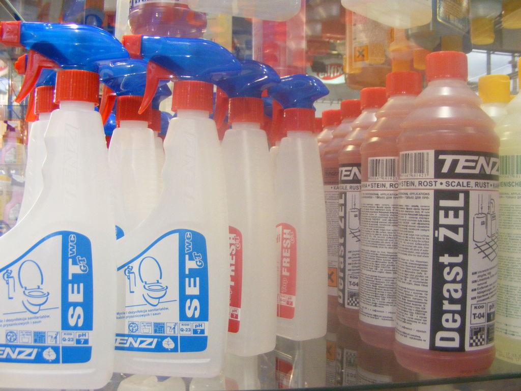Rząd butelek z płynami do czyszczenia i dezynfekcji toalet oraz preparatami do usuwania kamienia i rdzy, ustawione na półce sklepowej, oświetlone jasnym światłem.