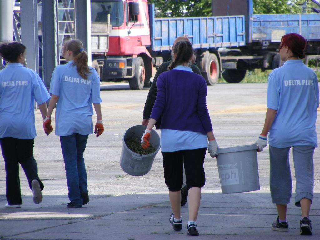Grupa osób w firmowych koszulkach Delta Plus niesie wiadra z resztkami roślinnymi na tle samochodów ciężarowych.