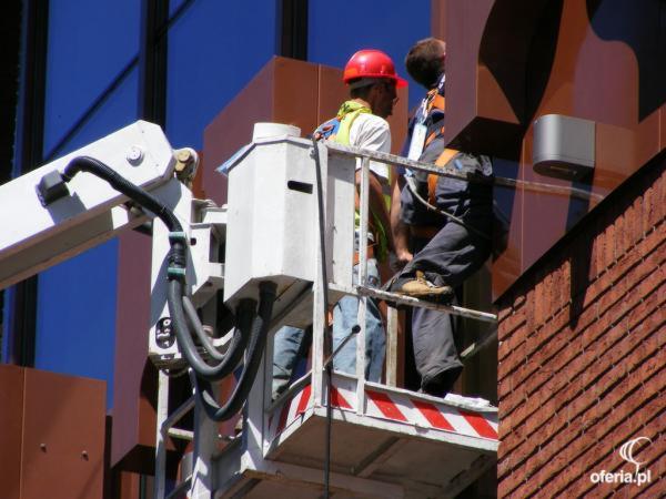 Dwóch pracowników na podnośniku koszowym, jeden w czerwonym kasku, drugi w uprzęży, pracujących na wysokości przy brązowej fasadzie budynku z elementami dekoracyjnymi i oknami.