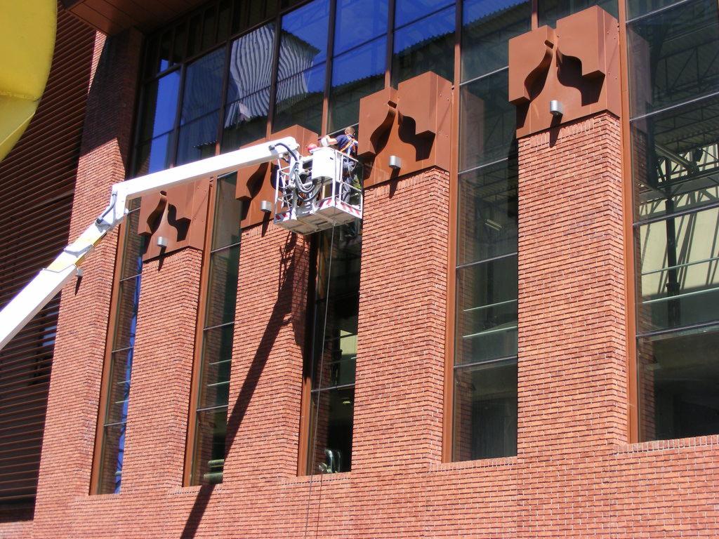 Pracownicy w koszu podnośnika teleskopowego myją wysokie okna budynku z cegły z dekoracyjnymi metalowymi elementami.