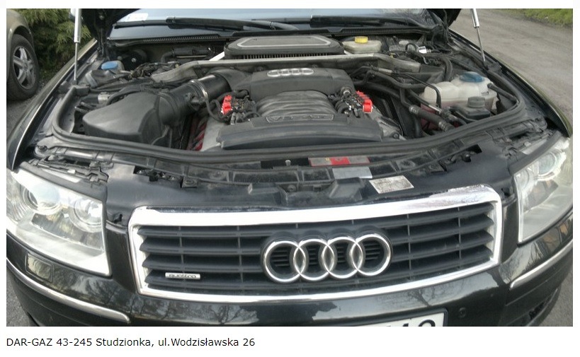 Czarny samochód marki Audi z otwartą maską, widoczny silnik V8 z czerwonymi elementami instalacji, na pierwszym planie grill z logo producenta.