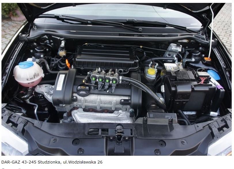 Silnik samochodu z zamontowaną instalacją LPG, widoczne wtryskiwacze gazu z zielonymi końcówkami, zbiornik płynu chłodniczego z niebieską nakrętką, czarny akumulator, otwarty przedni panel...