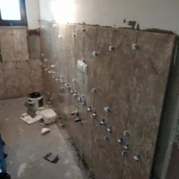 Układanie beżowych płytek na ścianie z użyciem klipsów poziomujących. Widoczne narzędzia i etap prac w łazience z oknem. Częściowo wykończone ściany.