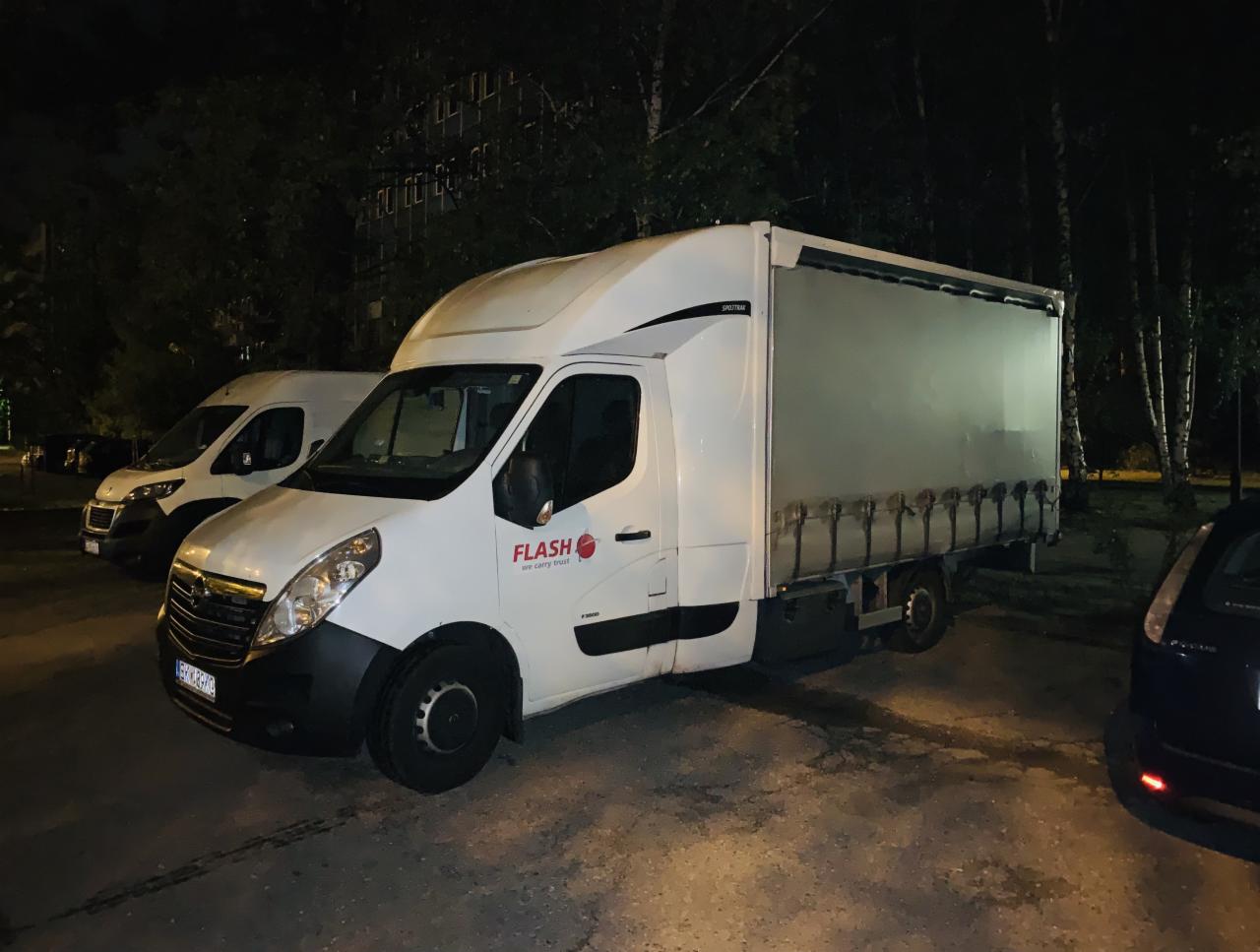 Biały samochód dostawczy marki Opel z logo firmy Flash zaparkowany na zewnątrz w nocy, widoczny drugi bus w tle.