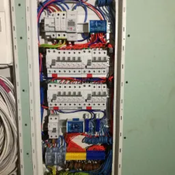 Rozdzielnica elektryczna z zainstalowanymi zabezpieczeniami, przewodami i listwami zaciskowymi, zamontowana w ścianie z płyt gipsowo-kartonowych, widoczne kable w kolorach niebieskim, czerwonym...