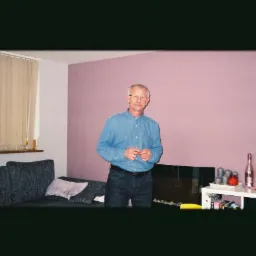 Mężczyzna w okularach, stojący w pokoju z fioletową ścianą, szarą kanapą i białym stolikiem z lampką, ramką ze zdjęciem i dekoracjami.