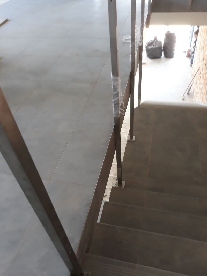 Metalowa balustrada schodowa z rur o kwadratowym przekroju, częściowo owiniętych folią ochronną, widok z góry na schody wyłożone płytkami.