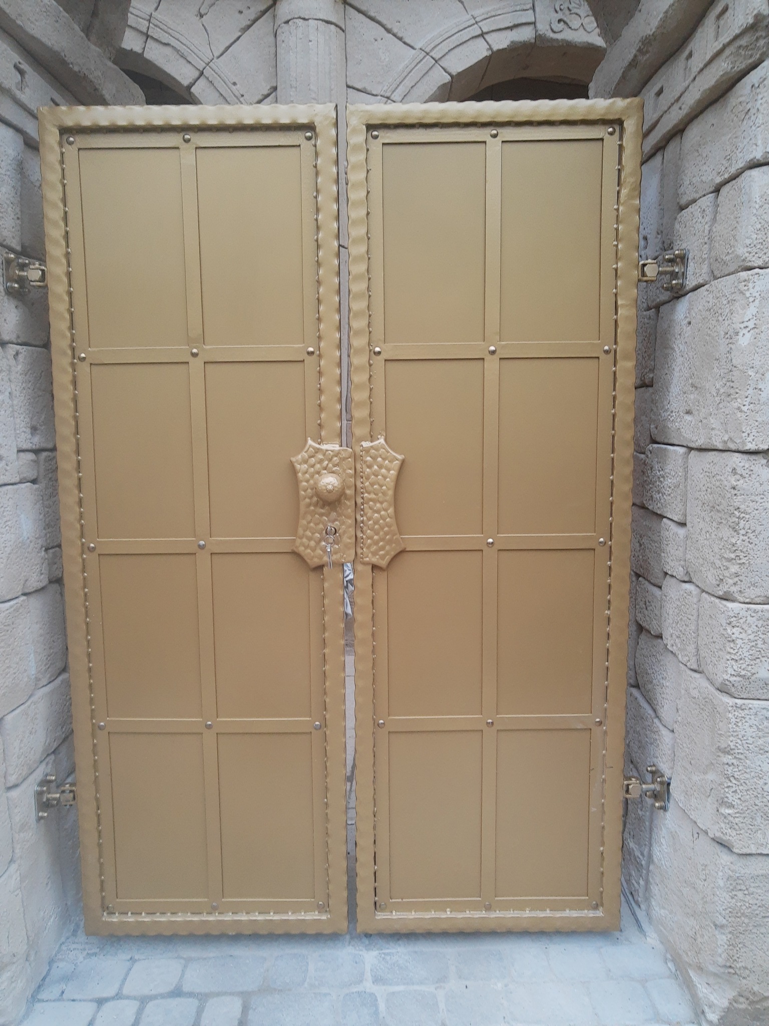 Złota, metalowa brama dwuskrzydłowa z ozdobnymi detalami, zamontowana w kamiennym murze, widoczne zawiasy i klucz w zamku.