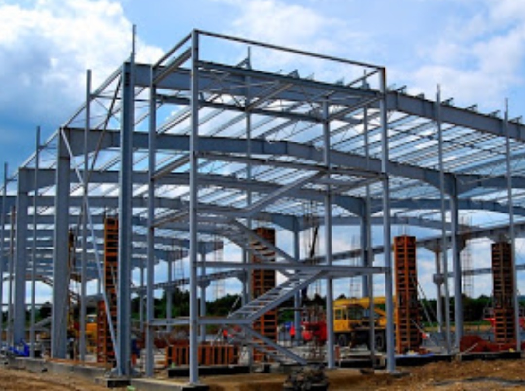 Wznoszenie szkieletu stalowego konstrukcji budynku przemysłowego, widoczne elementy nośne, schody i pomarańczowe szalunki, na tle błękitnego nieba.