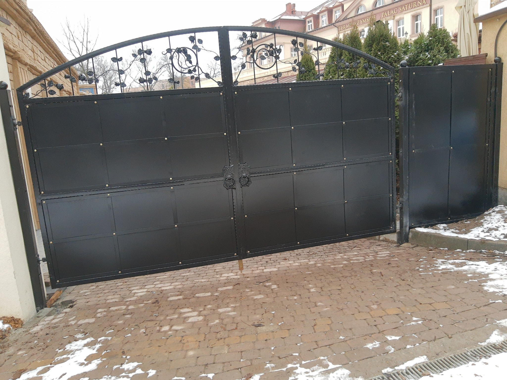 Czarna, metalowa brama wjazdowa z ozdobnymi elementami roślinnymi w górnej części, zamontowana na posesji z kostki brukowej.
