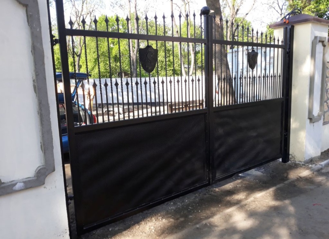 Czarna, metalowa brama wjazdowa z ozdobnymi grotami i tarczami herbowymi, zamontowana między dwoma murowanymi słupkami.