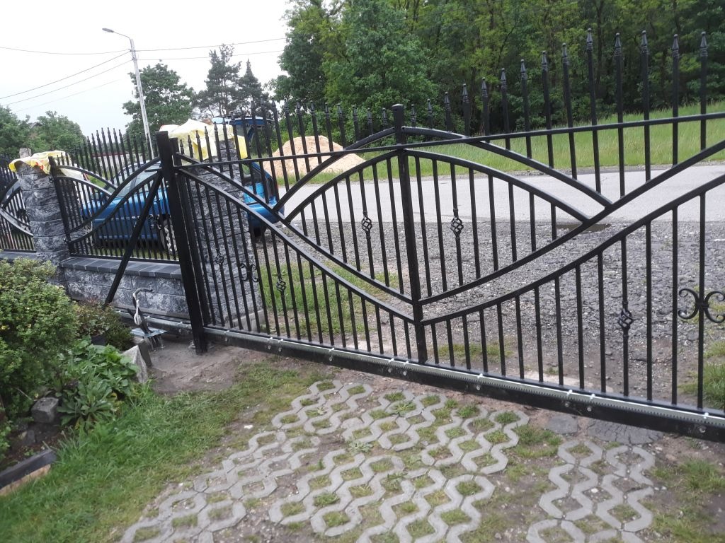 Czarna, metalowa brama przesuwna z dekoracyjnymi elementami, zamontowana na posesji z widocznym fragmentem podjazdu z kostki brukowej i zielenią w tle.