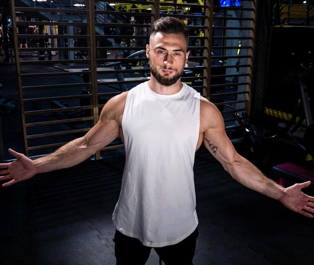 Mężczyzna w białym tank topie prezentuje sylwetkę na tle siłowni, z widocznym tatuażem na ramieniu, stojąc z rozłożonymi rękami.