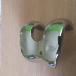Metalowy uchwyt w kształcie litery C, prawdopodobnie po procesie obróbki powierzchni, z widocznym odbiciem otoczenia na gładkiej powierzchni, leżący na drewnianej powierzchni.