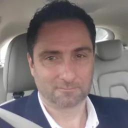 Portret mężczyzny w samochodzie, ubrany w marynarkę i koszulę, z zapiętym pasem bezpieczeństwa, uśmiechający się lekko.