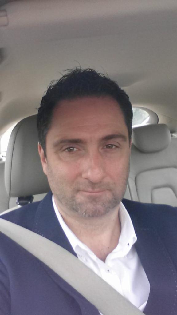 Portret mężczyzny w samochodzie, ubrany w marynarkę i koszulę, z zapiętym pasem bezpieczeństwa, uśmiechający się lekko.