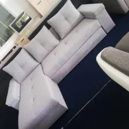 Jasnoszary narożnik modułowy z poduszkami na granatowym dywanie, widok z góry pod kątem. Fragment fotela z białym podłokietnikiem w kadrze.