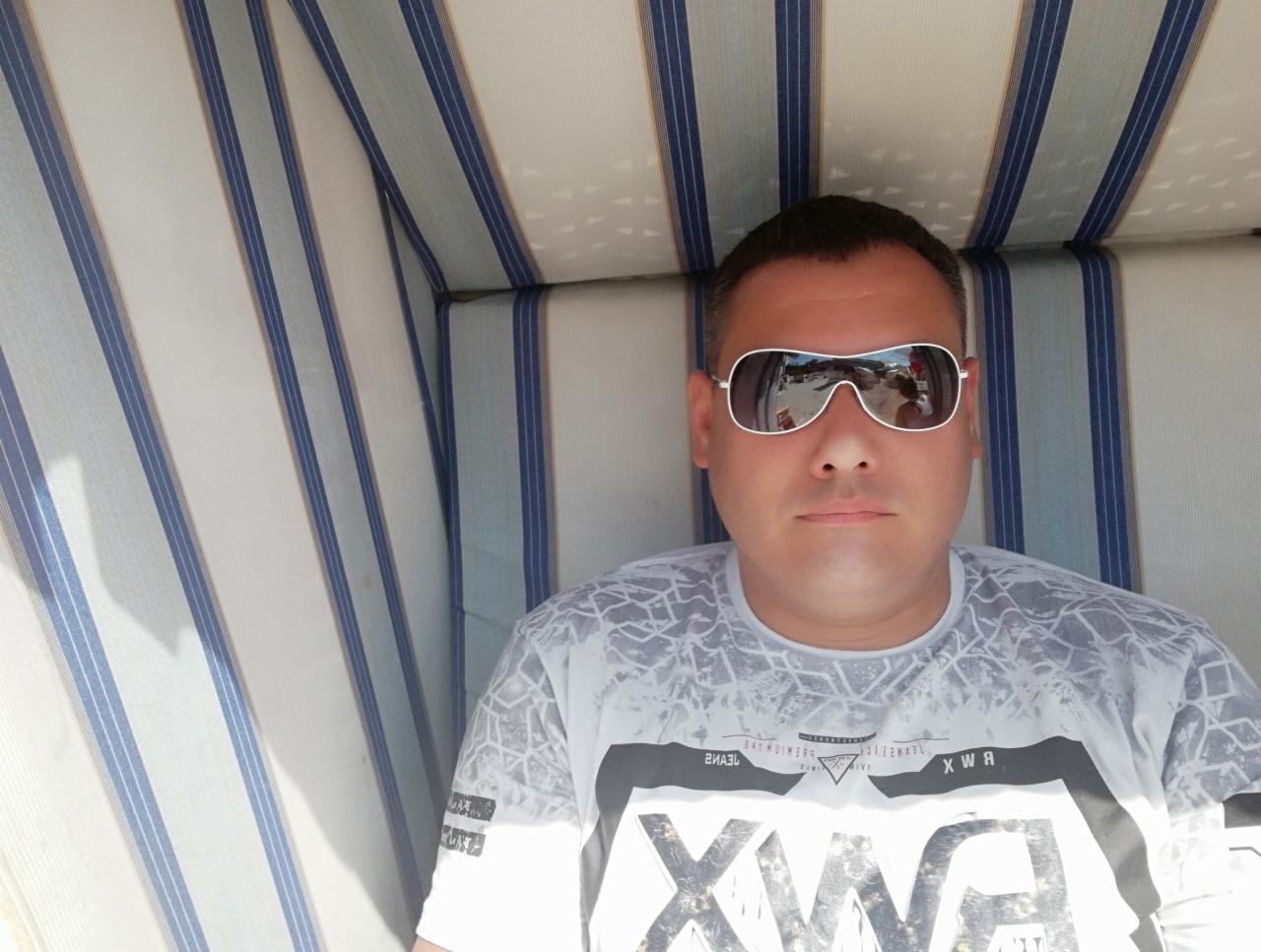 Mężczyzna w okularach przeciwsłonecznych siedzi w wiklinowym koszu plażowym z niebiesko-białymi pasami. W okularach odbija się krajobraz.