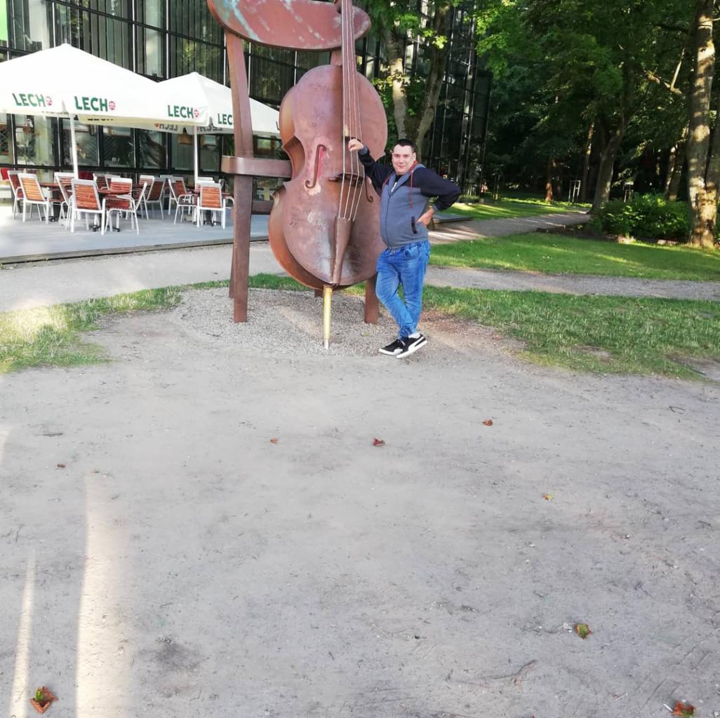 Mężczyzna opiera się o rdzewiejącą, metalową rzeźbę kontrabasu w parku, w tle budynek z przeszkloną fasadą i ogródek restauracyjny pod parasolami.