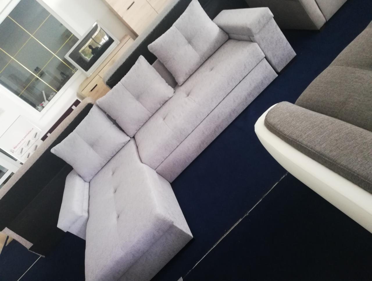 Jasnoszary narożnik modułowy z poduszkami na granatowym dywanie, widok z góry pod kątem. Fragment fotela z białym podłokietnikiem w kadrze.