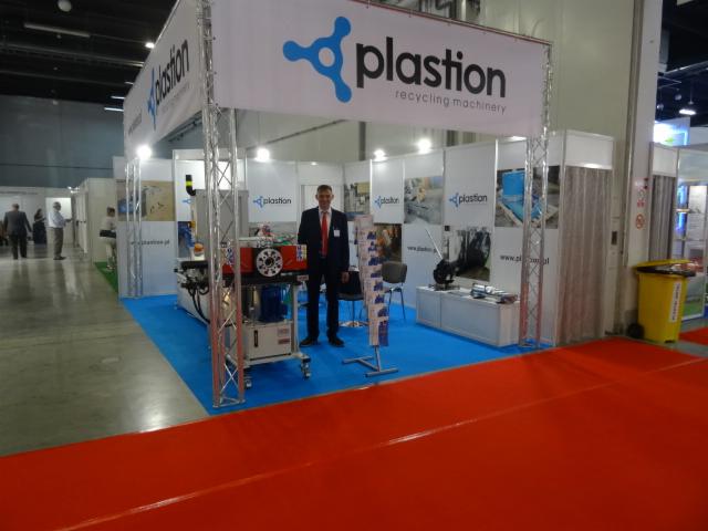 Stoisko targowe firmy zajmującej się maszynami do recyklingu z widocznym pracownikiem w garniturze i fragmentem maszyny na pierwszym planie.