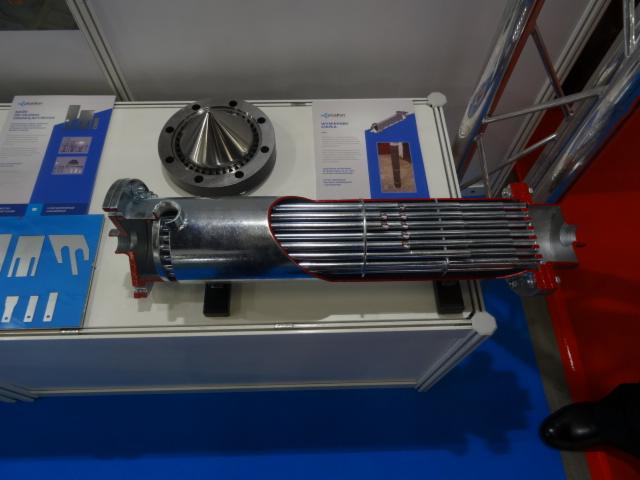 Przekrój poprzeczny wymiennika ciepła z rurkami, obok kołnierz i materiały informacyjne na stoisku targowym.