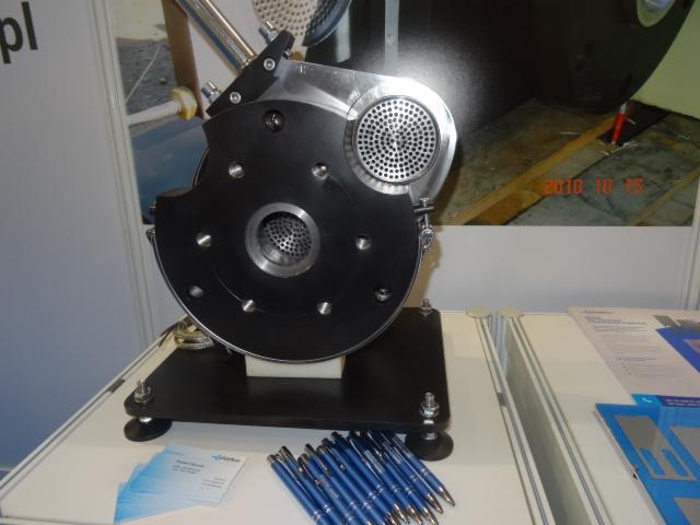 Prezentacja fragmentu maszyny z perforowanymi elementami na białym stole, obok wizytówka i zestaw niebieskich długopisów.