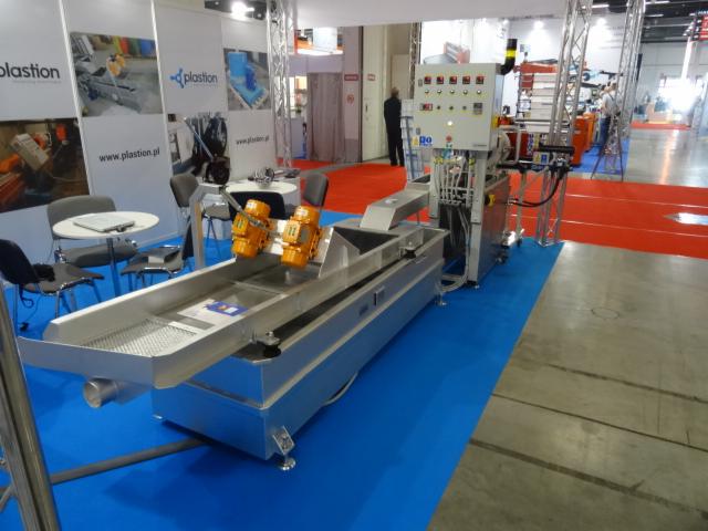 Srebrna maszyna przemysłowa z dwoma pomarańczowymi silnikami na targach, widoczny panel sterowania i rury, na podłodze niebieska wykładzina.