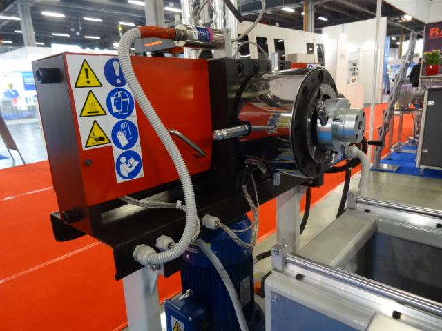 Czerwona maszyna przemysłowa z chromowanym elementem obrotowym, oznaczona naklejkami ostrzegawczymi BHP, na tle hali targowej.