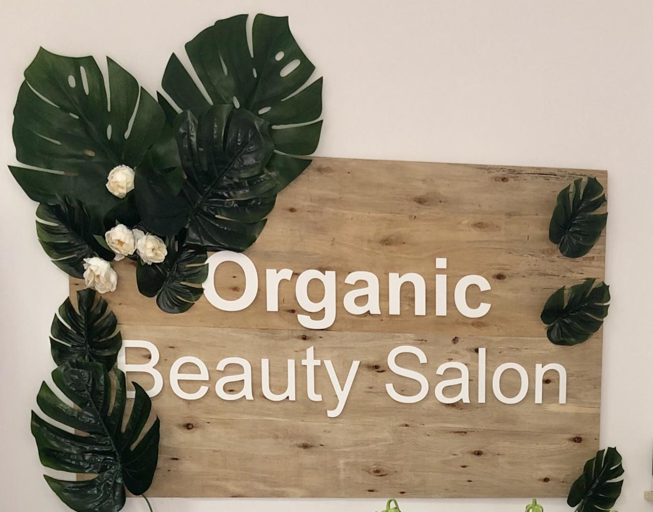 Drewniana tablica z napisem 'Organic Beauty Salon' ozdobiona liśćmi monstery i białymi kwiatami na jasnej ścianie.